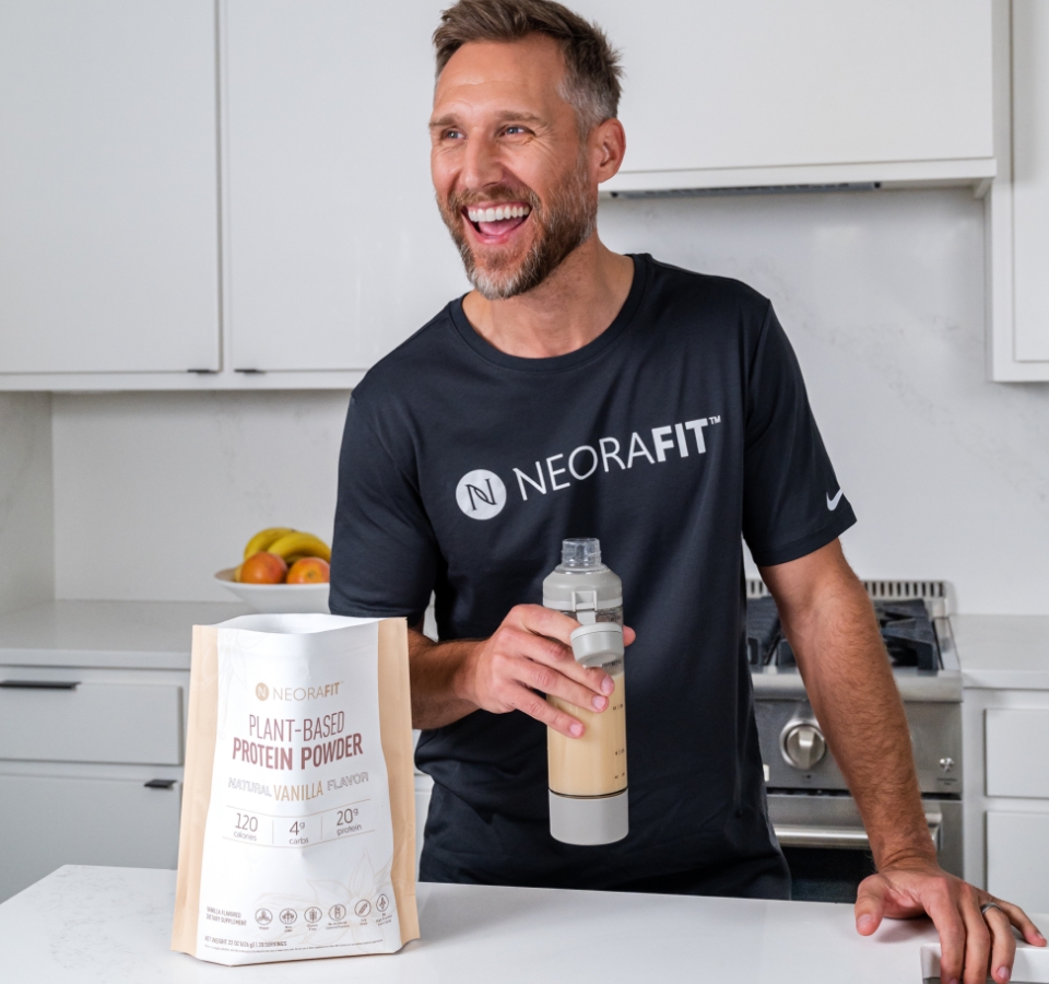 A man wearing a black NeoraFit shirt next to a kitchen counter holding up a glass of water with a bag of Neora's Plant-Based Protein Powder in front of him.